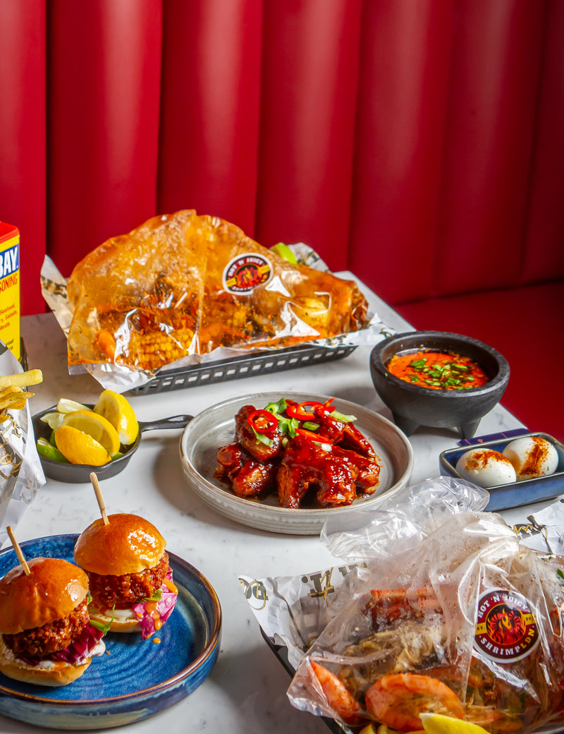 Table spread featuring sticky BBQ wings, boiled egg sprinkled with Old Bay, shrimp sliders, lemon slices, and a fresh seafood boil bag with sauce.